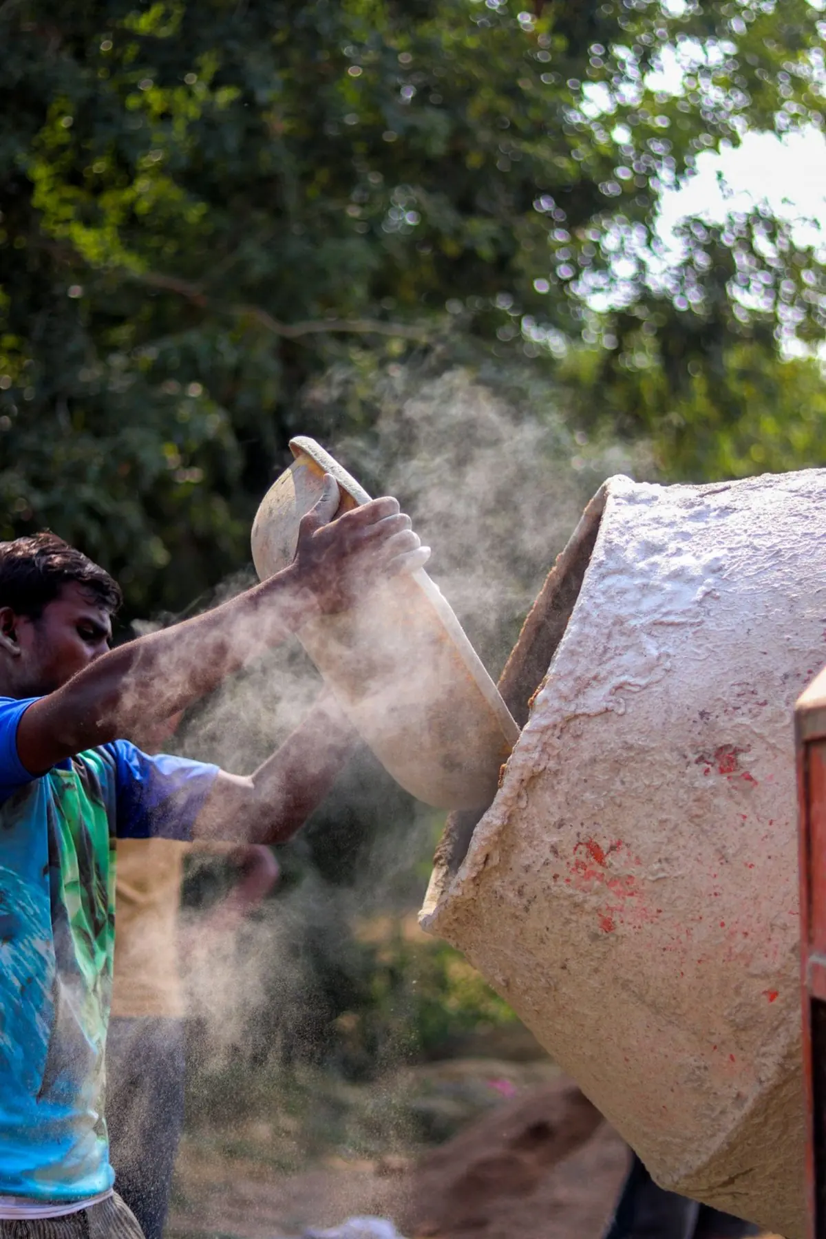 A worker tipping a pan of powder into a mixer on site, dust hanging in the air around him