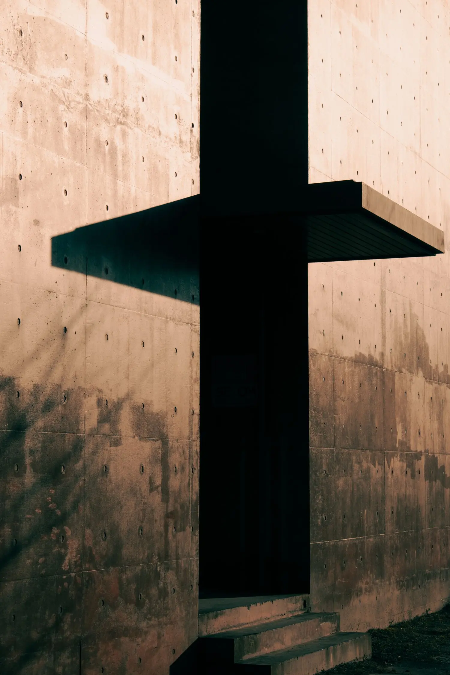 Raking light across a board-marked concrete wall, the tie holes of the formwork still visible, beside the deep shadow of a recessed entrance